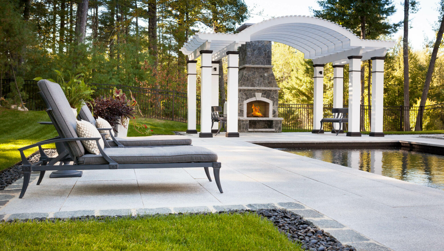 Chairs by the inground pool with pergola & outdoor fireplace in the background. Residential Landscape Design & Build Project.