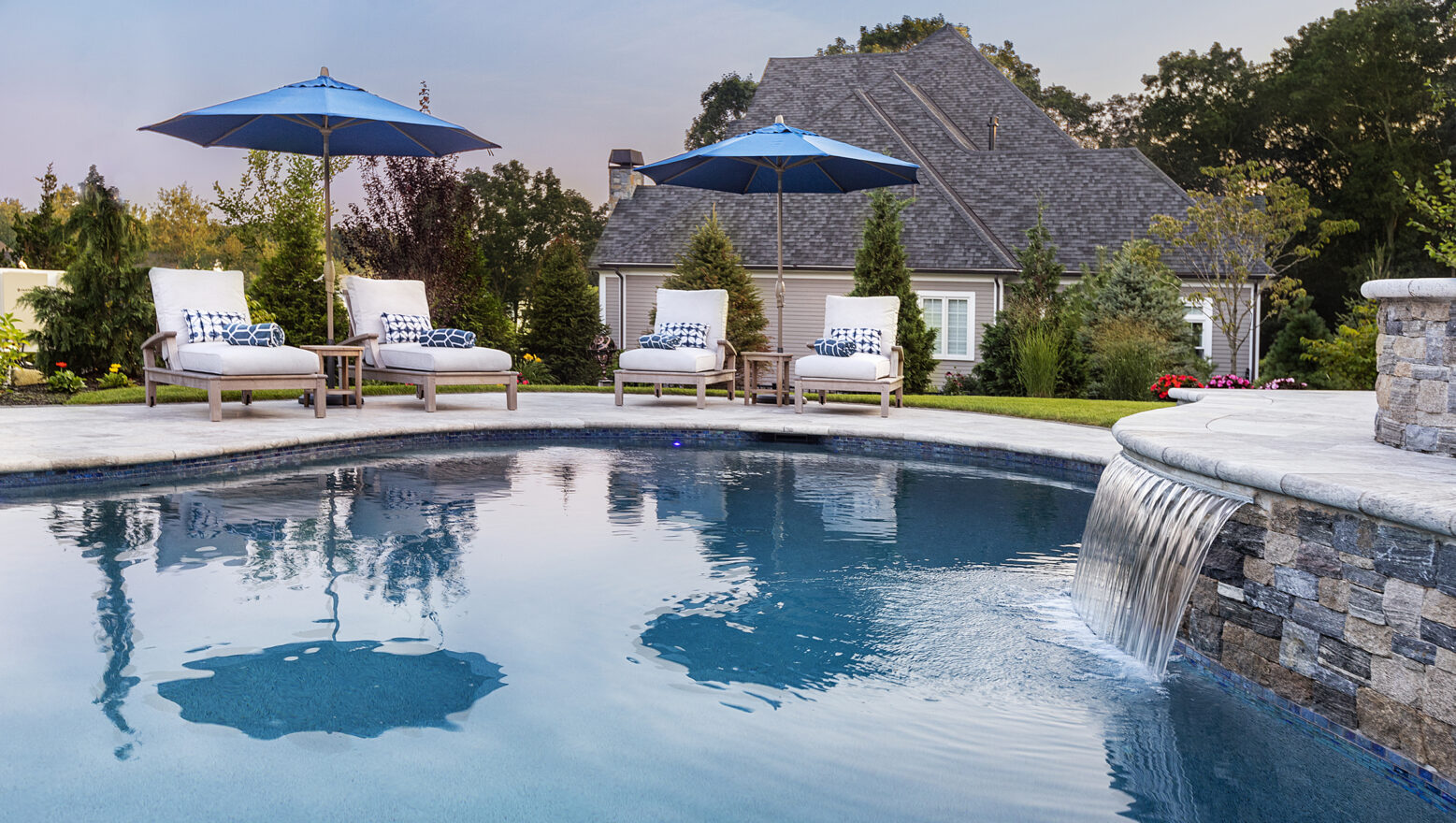 Pool chairs and water feature on a travertine pool deck built by Dex by Terra in Norfolk, Massachusetts.