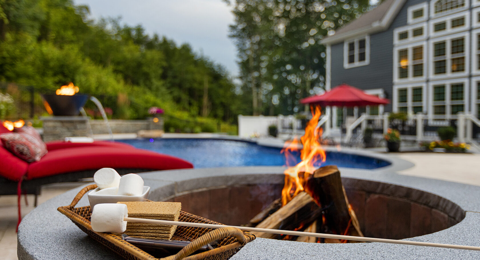 S'more kit by a wood-burning fire pit. Pool, fire bowl, and home in background. Dex by Terra Hardscaping in Sterling, MA.
