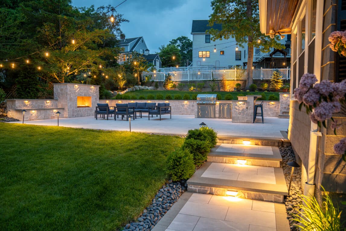 A natural stone paver walkway leads to a backyard patio with outdoor gas fireplace, and outdoor kitchen, illuminated by string lights and landscape lighting in Needham, MA.