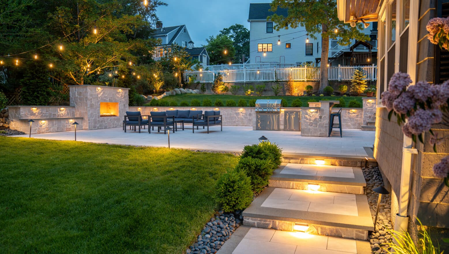 A natural stone paver walkway leads to a backyard patio with outdoor gas fireplace, and outdoor kitchen, illuminated by string lights and landscape lighting in Needham, MA.
