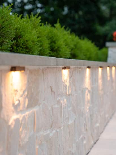 Stonewall with shrubs and low voltage outdoor landscape lighting in a Needham, MA backyard patio.